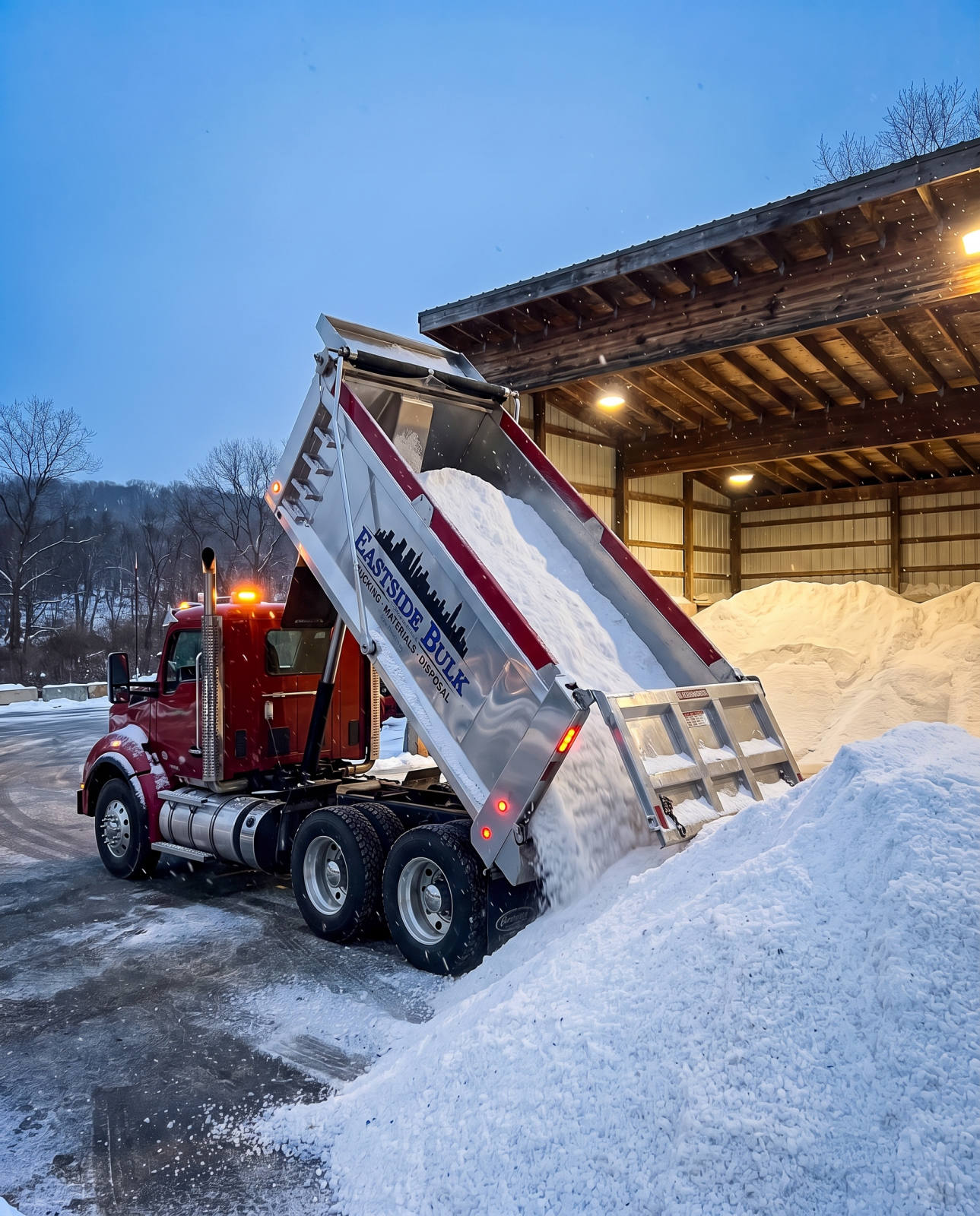 An Eastside Bulk dump truck tipping a load of white rock salt beside the salt shed on a snowy winter evening