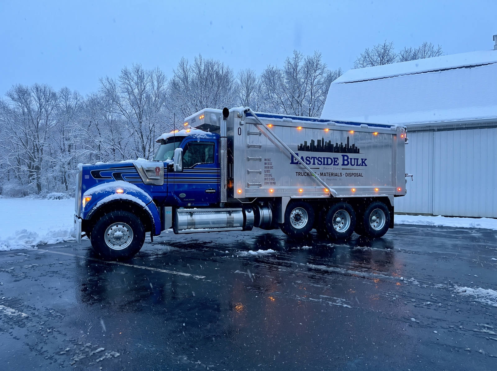 A blue Eastside Bulk tri-axle dump truck in a snowy yard beside a barn